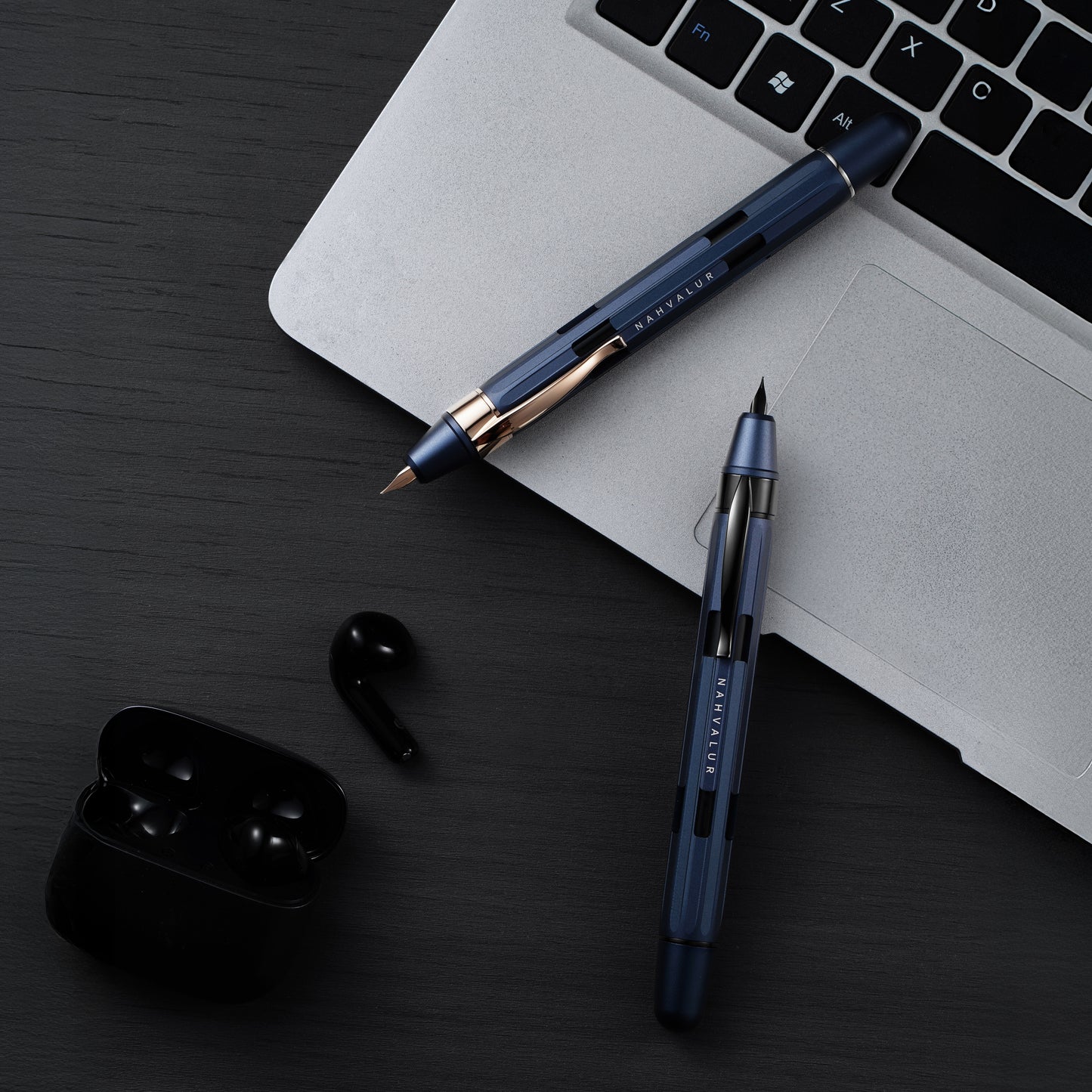 Two blue pens on a dark surface with a laptop and black earbuds.