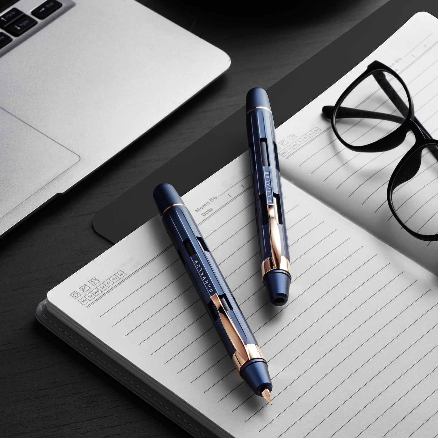 Two pens on a notebook with a laptop and glasses in the background