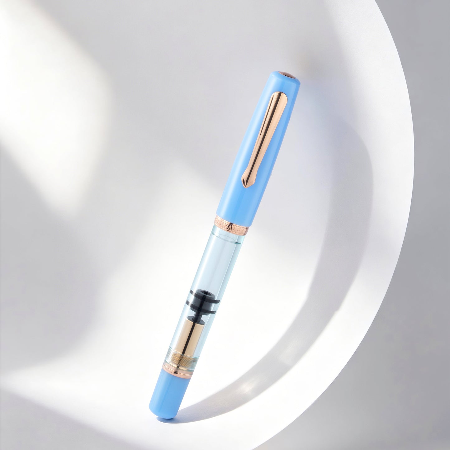 
                  
                    A light blue and clear glass pen with rose gold accents, resting diagonally on a white curved surface with soft shadows.
                  
                