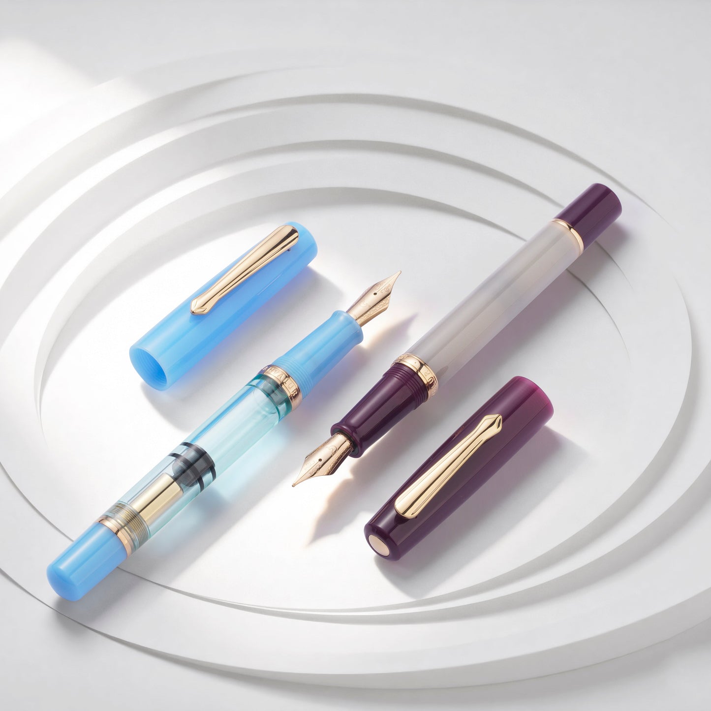 
                  
                    Four fountain pens arranged on a white circular pedestal. The set includes two blue pens, one translucent gray pen with a dark purple cap, and one dark purple pen with a gold nib and cap.
                  
                