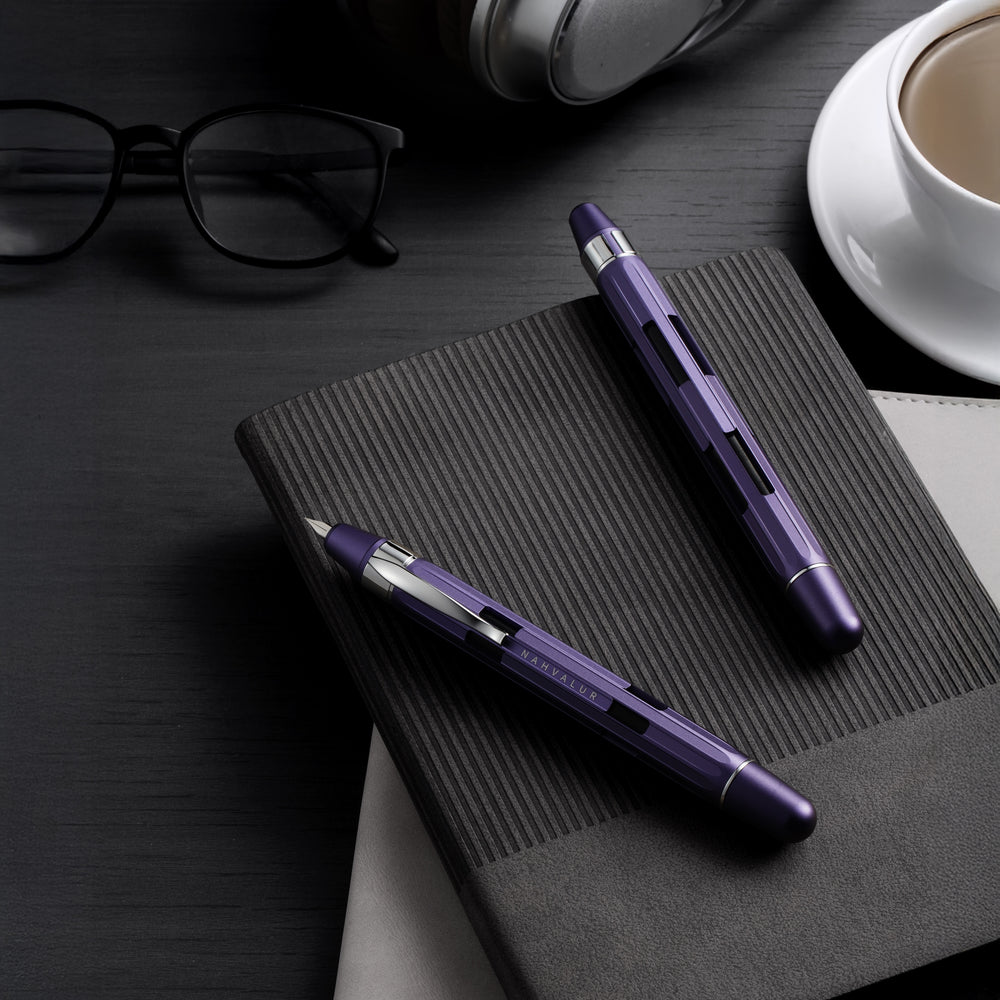 Purple pen and black notebook on a dark surface with glasses and a cup in the background