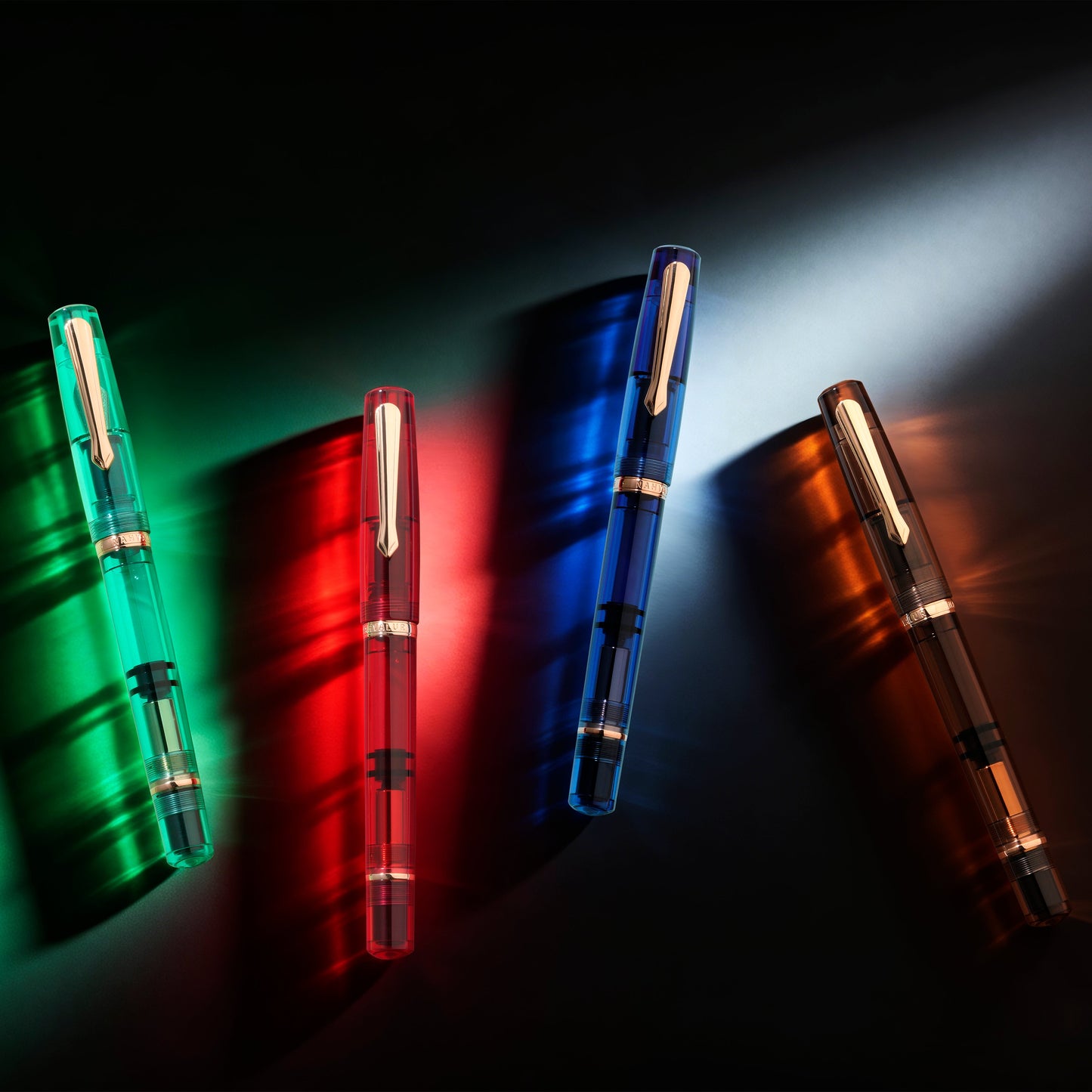 
                  
                    Four transparent fountain pens in green, red, blue, and orange, arranged diagonally on a dark background with dramatic colored lighting and shadows.
                  
                