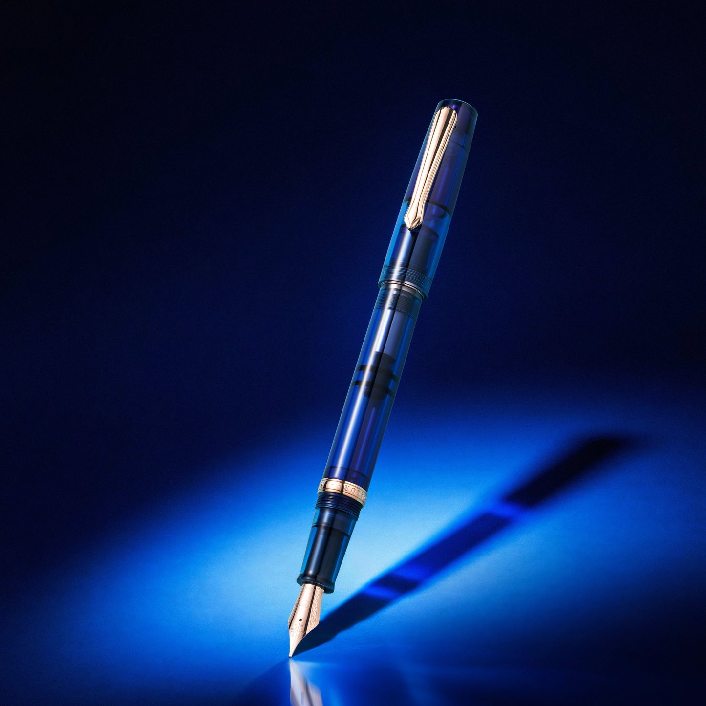 
                  
                    A sleek transparent blue fountain pen with silver metal accents, standing diagonally on a reflective dark surface under dramatic blue lighting.
                  
                