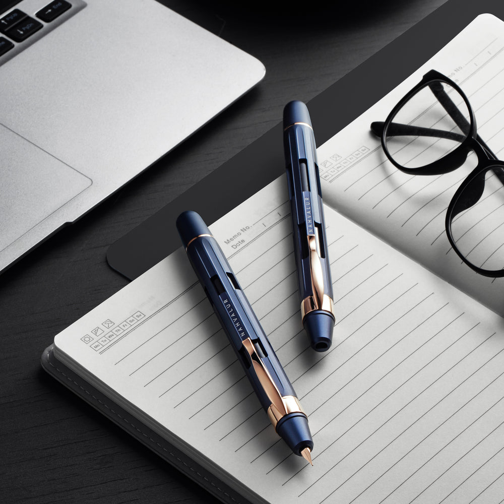 Two pens on a notebook with a laptop and glasses in the background